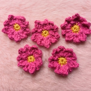 Könnte beinhalten: Fünf handgehäkelte Blumen in leuchtendem Fuchsia-Rosa mit gelben Zentren. Sie liegen auf einer weichen, rosa strukturierten Oberfläche. Die Nahaufnahme zeigt die detaillierte Häkelarbeit und den Farbkontrast.