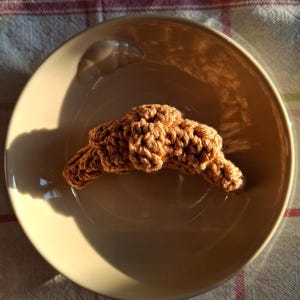 Può includere: Un croissant marrone all'uncinetto è appoggiato su un piatto beige. Il piatto è su una tovaglia a scacchi. Il croissant è fatto di filato strutturato e ha un aspetto annodato.