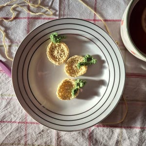 May include: Three crocheted yellow and green pineapple-shaped coasters sit on a white plate with black stripes. The coasters have a circular design with green leafy tops. A cup of tea is visible in the background.