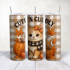 May include: A tumbler with a fall theme. The design features a cartoon cat with an orange bow and a black and white scarf, pumpkins, maple leaves, and the words "Cutin' N Cuddly" on a tan and white plaid background. Includes a straw.