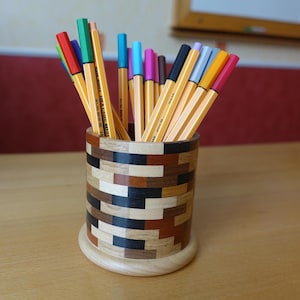 Peut inclure: Un porte-crayons cylindrique en bois présentant un motif en mosaïque composé de blocs de bois clairs, moyens et foncés. Le support est rempli d'une collection de stylos feutres multicolores dans des tons de rouge, vert, bleu, jaune, rose et noir.