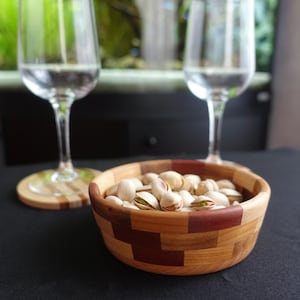 Peut inclure: Un bol rond en bois présentant un motif en mosaïque composé de segments de bois clair et brun foncé, rempli de pistaches. Le bol est posé sur une surface noire à côté de deux verres à vin transparents et d'un dessous de verre assorti.