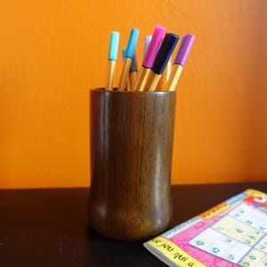 Peut inclure: Un porte-crayons cylindrique en bois brun foncé contenant plusieurs feutres multicolores avec des corps rayés jaunes. Les stylos présentent des capuchons dans des tons bleu clair, rose, bleu foncé et violet, disposés verticalement.
