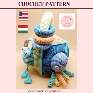 Può includere: Un giocattolo all'uncinetto di colore blu, verde e arancione. Il giocattolo ha la forma di un cubo con un manico in cima e quattro anelli che possono essere impilati sul manico. Il giocattolo ha anche un viso all'uncinetto con occhi e bocca. Il giocattolo è realizzato da "Smoozy Crochet Patterns".