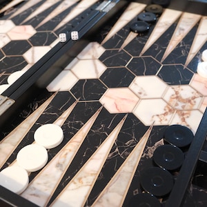 May include: A backgammon board with a black frame, featuring a black and marble-patterned playing surface. White and black game pieces are arranged on the board, along with dice. The board is open, ready for play.