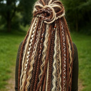 Op de afbeelding: Lang, gestyled dreadlocks in bruin- en blondtinten, met vlechten, wikkelingen en decoratieve elementen. De persoon draagt een donkergroene top. De achtergrond is een vage groene landschap.