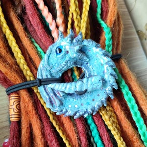 Könnte beinhalten: Nahaufnahme eines bunten Sets synthetischer Dreadlocks mit einem blauen, drachenförmigen Haargummi und einem silbernen Pentagramm-Anhänger. Die Dreadlocks sind in Rot-, Orange-, Gelb-, Grün- und Blautönen gehalten.