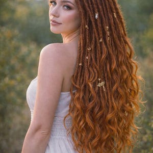 May include: A woman with long, auburn dreadlocks decorated with gold hair accessories. She wears a white dress and looks over her shoulder. The dreadlocks flow down her back, with a natural outdoor background.
