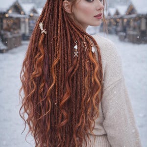 May include: A woman with long, auburn dreadlocks adorned with decorative hair jewelry. The dreadlocks cascade down the back, with some sections styled into loose waves. She wears a cream-colored sweater, and the background shows a snowy, winter scene.