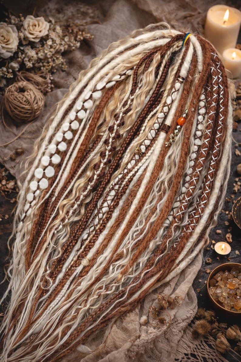 Op de afbeelding: Een verzameling bruine, witte en blonde dreadlocks, sommige versierd met decoratieve stiksels en kralen. De dreadlocks zijn in een waaier gerangschikt en tonen verschillende texturen en stijlen. De algehele esthetiek is boheems en natuurlijk.