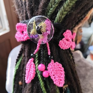 Puede incluir: Una liga para el pelo de crochet rosa con un dije de flor de resina. El dije presenta flores prensadas en tonos de morado, rosa y blanco.