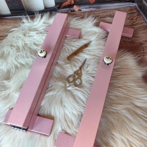 May include: Two pink wooden embroidery hoops with decorative knobs, a small wooden tool, and gold scissors rest on a fluffy white surface. The hoops are designed for holding fabric taut during needlework projects.