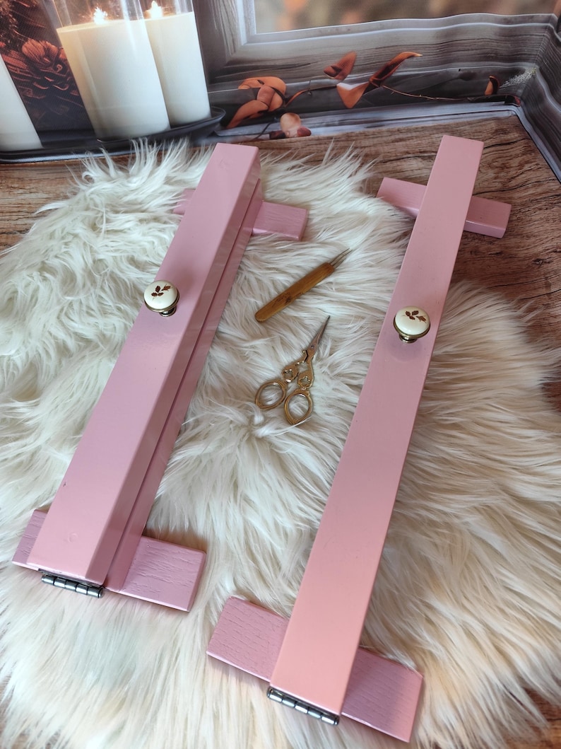 May include: Two pink wooden embroidery hoops with decorative knobs, a small wooden tool, and gold scissors rest on a fluffy white surface. The hoops are designed for holding fabric taut during needlework projects.