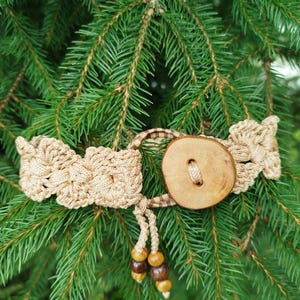 Puede incluir: Un cinturón de crochet beige con un botón de madera y cuentas de madera. El cinturón está en una rama de pino verde.