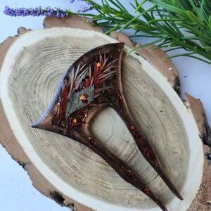 May include: A brown hair fork with a resin design featuring dried flowers and glitter. The fork has two prongs and is set on a wooden surface.