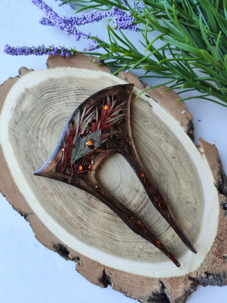 May include: A brown hair fork with a resin design featuring dried flowers and glitter. The fork has two prongs and is set on a wooden surface.