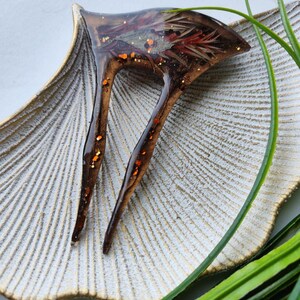 May include: A brown resin hair clip with two prongs. The clip has a floral design with red, orange, and gold glitter embedded in the resin.