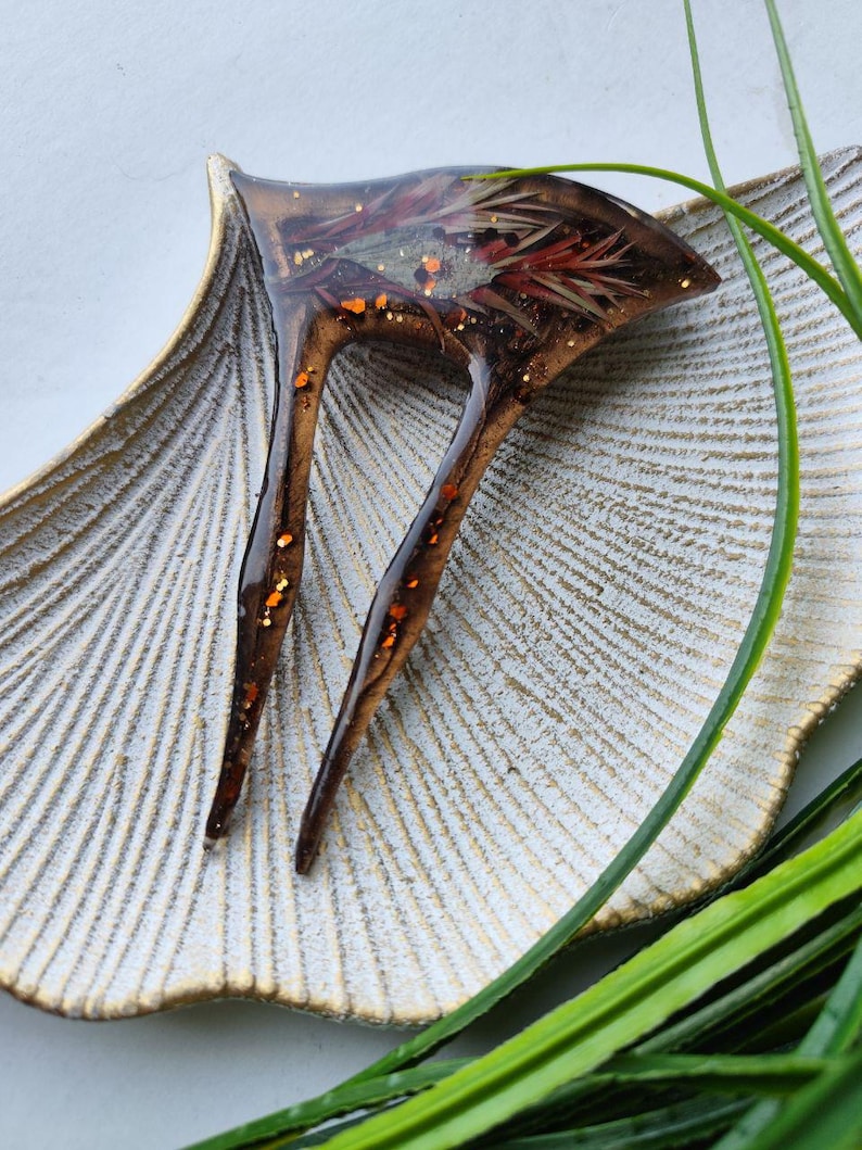 May include: A brown resin hair clip with two prongs. The clip has a floral design with red, orange, and gold glitter embedded in the resin.