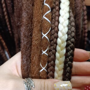 K&ouml;nnte beinhalten: Nahaufnahme von braunen, wei&szlig;en und schwarzen synthetischen Dreadlocks mit wei&szlig;en N&auml;hten. Die Dreadlocks werden in einer Hand gehalten, die einen silbernen Ring tr&auml;gt.