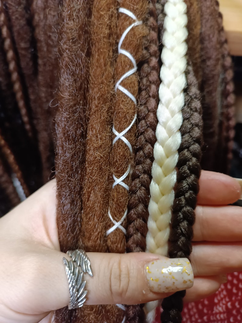 K&ouml;nnte beinhalten: Nahaufnahme von braunen, wei&szlig;en und schwarzen synthetischen Dreadlocks mit wei&szlig;en N&auml;hten. Die Dreadlocks werden in einer Hand gehalten, die einen silbernen Ring tr&auml;gt.