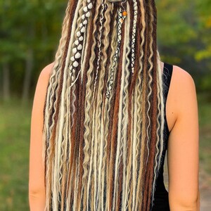 Op de afbeelding: Achteraanzicht van een persoon met lange, golvende dreadlocks in bruine, blonde en cr&egrave;metinten. Het haar is opgestoken in een knot op de kruin en versierd met kralen en bedels. De persoon draagt een zwarte top.