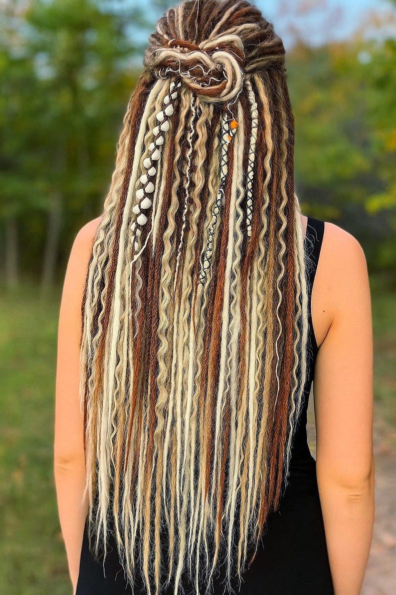 Op de afbeelding: Achteraanzicht van een persoon met lange, golvende dreadlocks in bruine, blonde en cr&egrave;metinten. Het haar is opgestoken in een knot op de kruin en versierd met kralen en bedels. De persoon draagt een zwarte top.