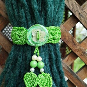 May include: A handmade, dark green scarf is adorned with a bright green crocheted band featuring a large, clear button with a floral design. A beaded tassel with green and white beads and crocheted leaf accents hangs below the button, adding a decorative touch.