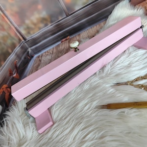 May include: A light pink knitting needle case with a cream-colored knob. The case is rectangular and has a hinged lid. The case is resting on a white fluffy surface. A wooden knitting needle and gold scissors are also visible.
