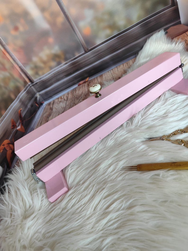 May include: A light pink knitting needle case with a cream-colored knob. The case is rectangular and has a hinged lid. The case is resting on a white fluffy surface. A wooden knitting needle and gold scissors are also visible.
