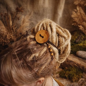 Puede incluir: Primer plano de un peinado con rastas rubias, adornado con un botón grande y redondo de madera y una cadena de pequeñas cuentas de color ámbar. El botón está sujeto a un cordón de cuero marrón, añadiendo una estética natural y bohemia.