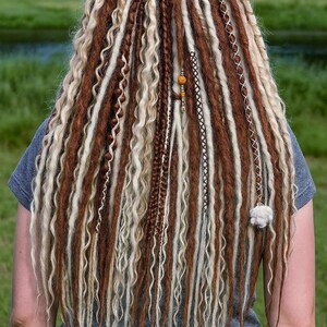 Op de afbeelding: Achterkant van een persoon met lange, bruine, blonde en witte dreadlocks. De dreadlocks zijn gestyled met verschillende vlechten en decoratieve elementen, waaronder een gele band en kralen. Het haar valt over de rug.