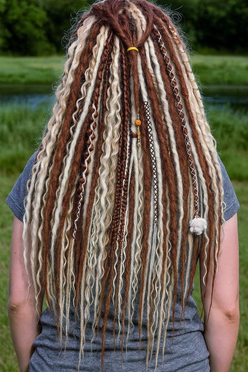 Op de afbeelding: Achterkant van een persoon met lange, bruine, blonde en witte dreadlocks. De dreadlocks zijn gestyled met verschillende vlechten en decoratieve elementen, waaronder een gele band en kralen. Het haar valt over de rug.