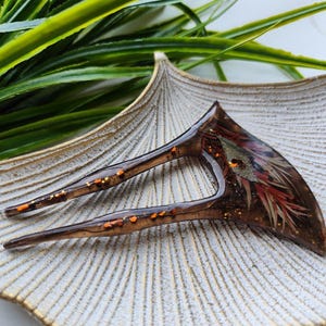 May include: A brown and orange hair fork with gold glitter embedded in the resin. The fork is shaped like a crescent moon and has two prongs.