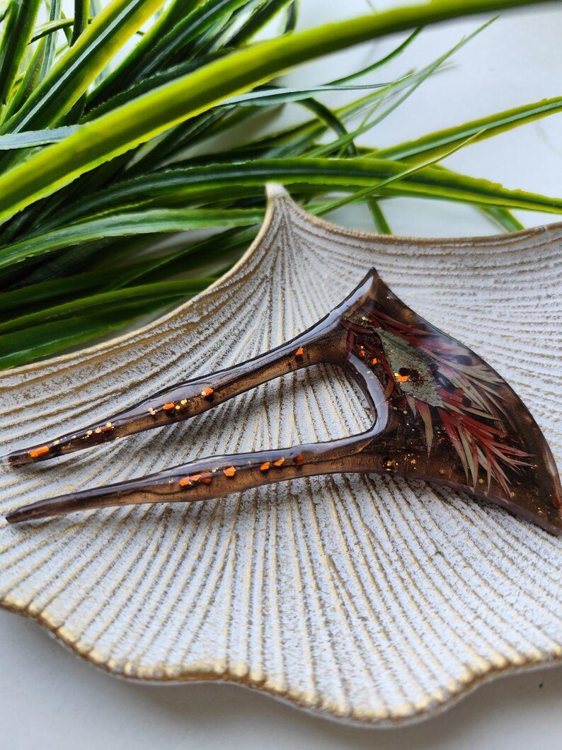 May include: A brown and orange hair fork with gold glitter embedded in the resin. The fork is shaped like a crescent moon and has two prongs.
