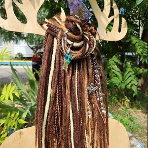 K&ouml;nnte beinhalten: Braune und wei&szlig;e synthetische Dreadlocks mit dekorativen Anh&auml;ngern, darunter ein Schmetterling, eine Blume und ein Kristall. Die Dreadlocks sind zu einem Knoten gebunden und an einem Holz-Geweih-Stirnband befestigt.