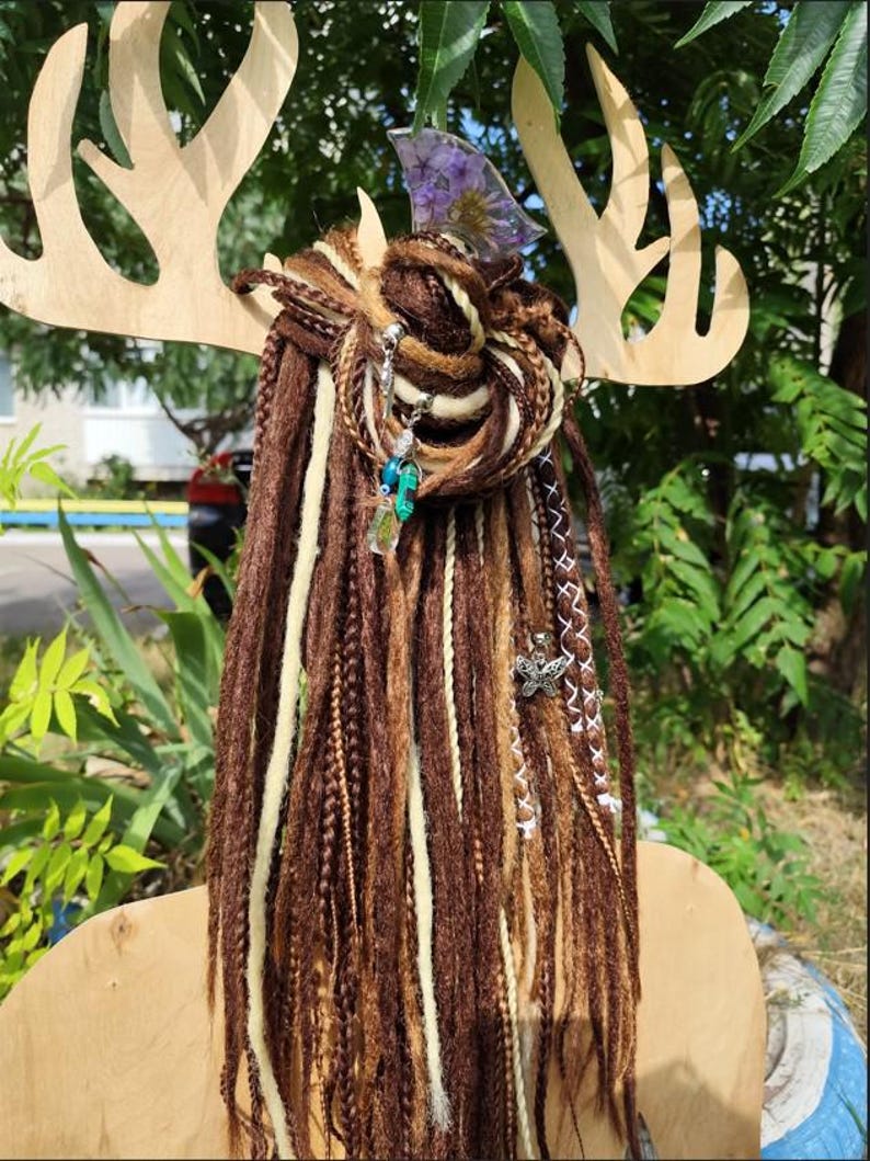 K&ouml;nnte beinhalten: Braune und wei&szlig;e synthetische Dreadlocks mit dekorativen Anh&auml;ngern, darunter ein Schmetterling, eine Blume und ein Kristall. Die Dreadlocks sind zu einem Knoten gebunden und an einem Holz-Geweih-Stirnband befestigt.