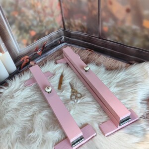 May include: Two pink wooden embroidery hoops with white knobs and silver hinges are displayed on a fluffy white rug. A small wooden tool and gold scissors are also visible, suggesting a crafting theme.