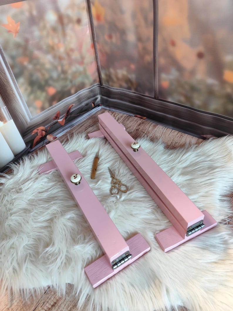 May include: Two pink wooden embroidery hoops with white knobs and silver hinges are displayed on a fluffy white rug. A small wooden tool and gold scissors are also visible, suggesting a crafting theme.