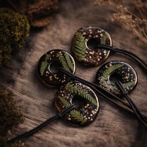 Puede incluir: Cuatro colgantes circulares con resina negra, hojas de helecho y motas doradas. Cada colgante tiene un agujero central y está ensartado en un cordón negro. Los colgantes descansan sobre una superficie marrón texturizada.