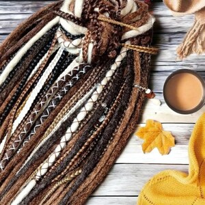 K&ouml;nnte beinhalten: Nahaufnahme eines B&uuml;ndels brauner, wei&szlig;er und schwarzer synthetischer Dreadlocks. Die Dreadlocks sind oben zusammengebunden und haben verschiedene Verzierungen, darunter Perlen und N&auml;hte.