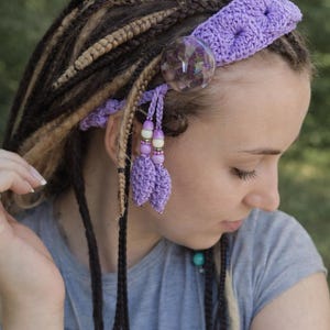 Puede incluir: Una diadema de ganchillo lila con un adorno redondo transparente y borlas colgantes. La diadema se usa con rastas. Las borlas tienen cuentas blancas y lilas. El tema general es bohemio.