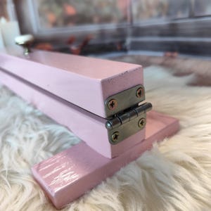 May include: A small, pink, wooden folding table with a silver hinge and screws. The table has a small, round, silver knob on the top. The table is sitting on a white, fluffy surface.