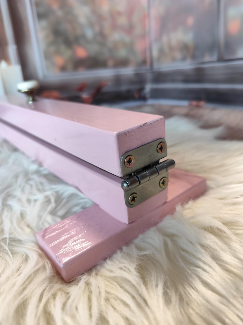 May include: A small, pink, wooden folding table with a silver hinge and screws. The table has a small, round, silver knob on the top. The table is sitting on a white, fluffy surface.