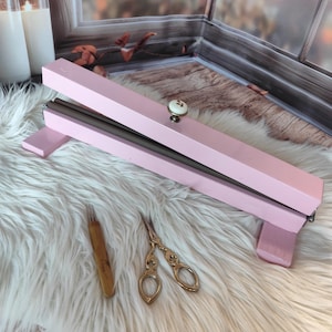 May include: A pink wooden embroidery frame with a decorative knob, a wooden needle tool, and gold-colored scissors. The frame is open, revealing a dark interior. The items rest on a white, fluffy surface.
