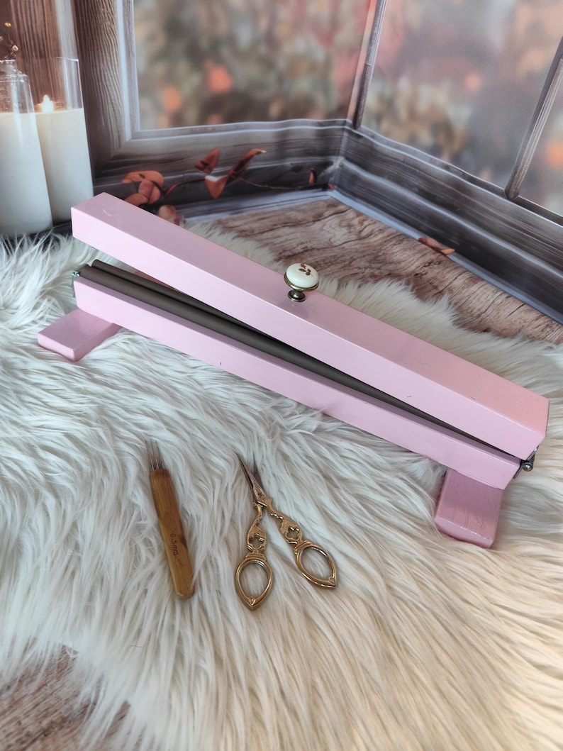 May include: A pink wooden embroidery frame with a decorative knob, a wooden needle tool, and gold-colored scissors. The frame is open, revealing a dark interior. The items rest on a white, fluffy surface.