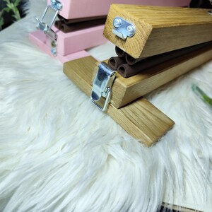 May include: Close-up of two wooden staplers. One is painted pink, the other is natural wood. Both have silver metal clasps and brown rubber components. The staplers are on a white, fluffy surface.