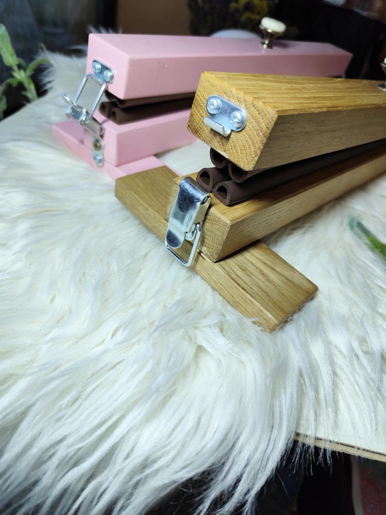 May include: Close-up of two wooden staplers. One is painted pink, the other is natural wood. Both have silver metal clasps and brown rubber components. The staplers are on a white, fluffy surface.