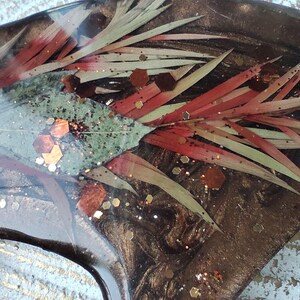 May include: Close-up of a resin art piece with a dark brown background. The piece features red and green leaves, and gold glitter.