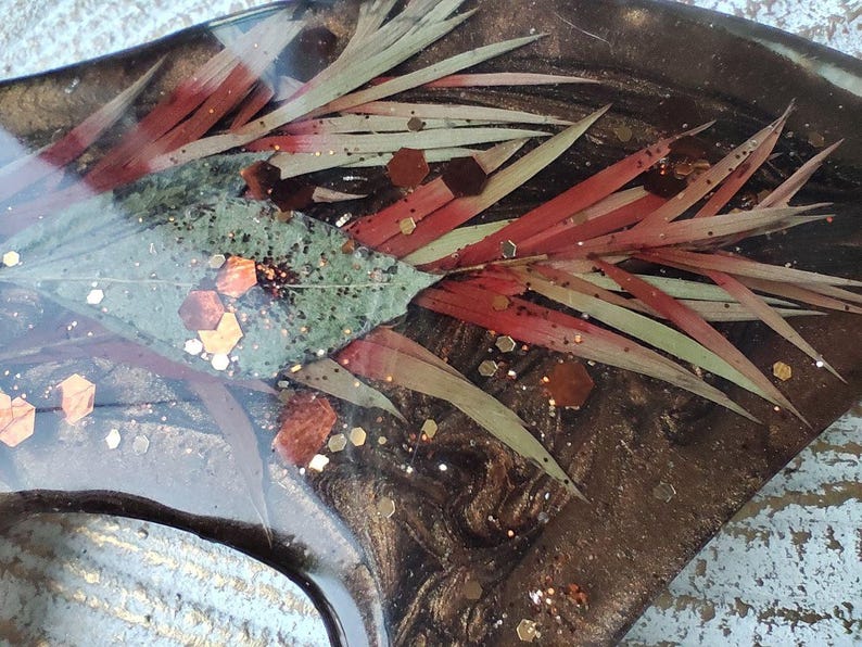 May include: Close-up of a resin art piece with a dark brown background. The piece features red and green leaves, and gold glitter.