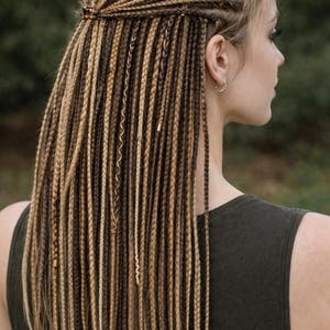 May include: A close-up of long, braided hair in shades of brown and blonde. The braids are styled with some pulled back and secured, while the rest cascade down the back. The person is wearing a dark green tank top.